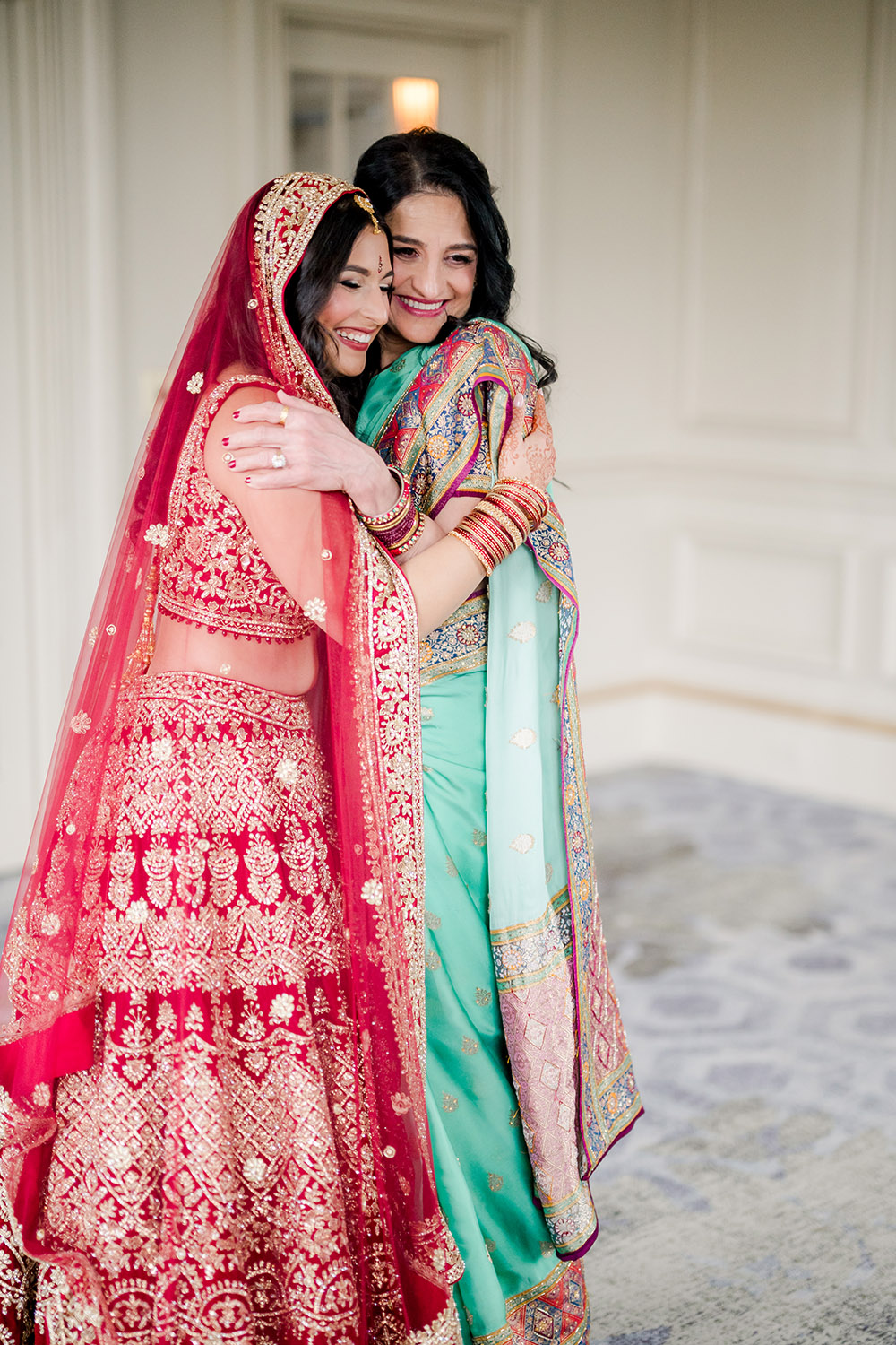 Bride in a red and gold embroidered lehenga warmly embraces a woman wearing a teal saree with colorful embroidered details. Both smile with eyes closed, sharing an emotional moment indoors before the ceremony.