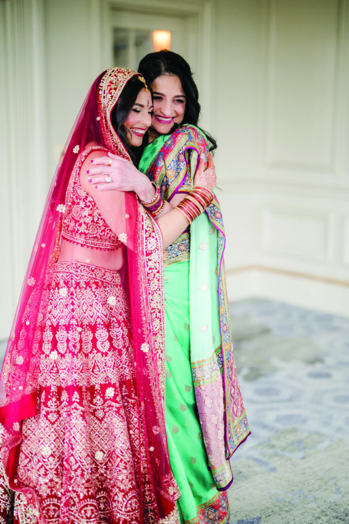 Bride in a red and gold embroidered lehenga warmly embraces a woman wearing a teal saree with colorful embroidered details. Both smile with eyes closed, sharing an emotional moment indoors before the ceremony.