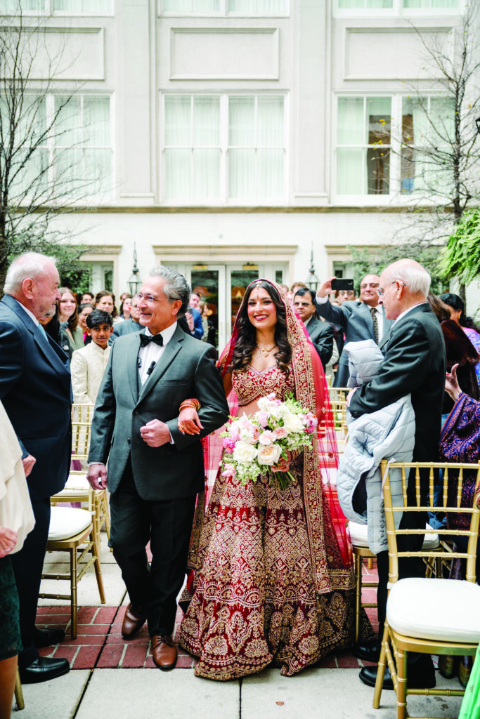Here’s a clear, detailed, and culturally accurate alt text for this image:

**Alt Text:**
“An Indian bride in a red and gold embroidered lehenga walks down the aisle with her father during an outdoor wedding ceremony. She carries a pastel bouquet and smiles as guests watch and take photos from both sides of the aisle.”
