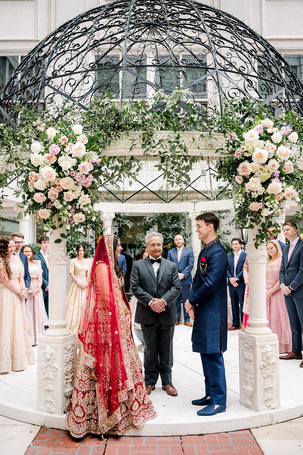 Bride and groom stand facing each other under an ornate floral mandap during their outdoor Indian wedding ceremony. The bride wears a red and gold embroidered lehenga with a matching veil, while the groom wears a navy blue sherwani. The officiant stands between them as their wedding party, dressed in coordinating pastel and navy attire, looks on.