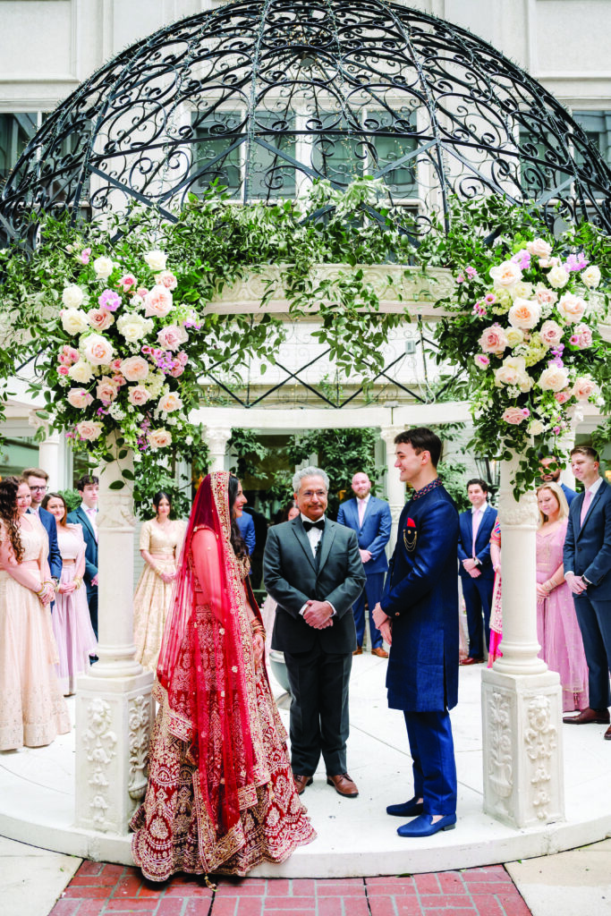 Here is a clear, detailed, and culturally accurate alt text for the image:

**Alt Text:**
“Bride and groom stand facing each other under an ornate floral mandap during their outdoor Indian wedding ceremony. The bride wears a red and gold embroidered lehenga with a matching veil, while the groom wears a navy blue sherwani. The officiant stands between them as their wedding party, dressed in coordinating pastel and navy attire, looks on.”

