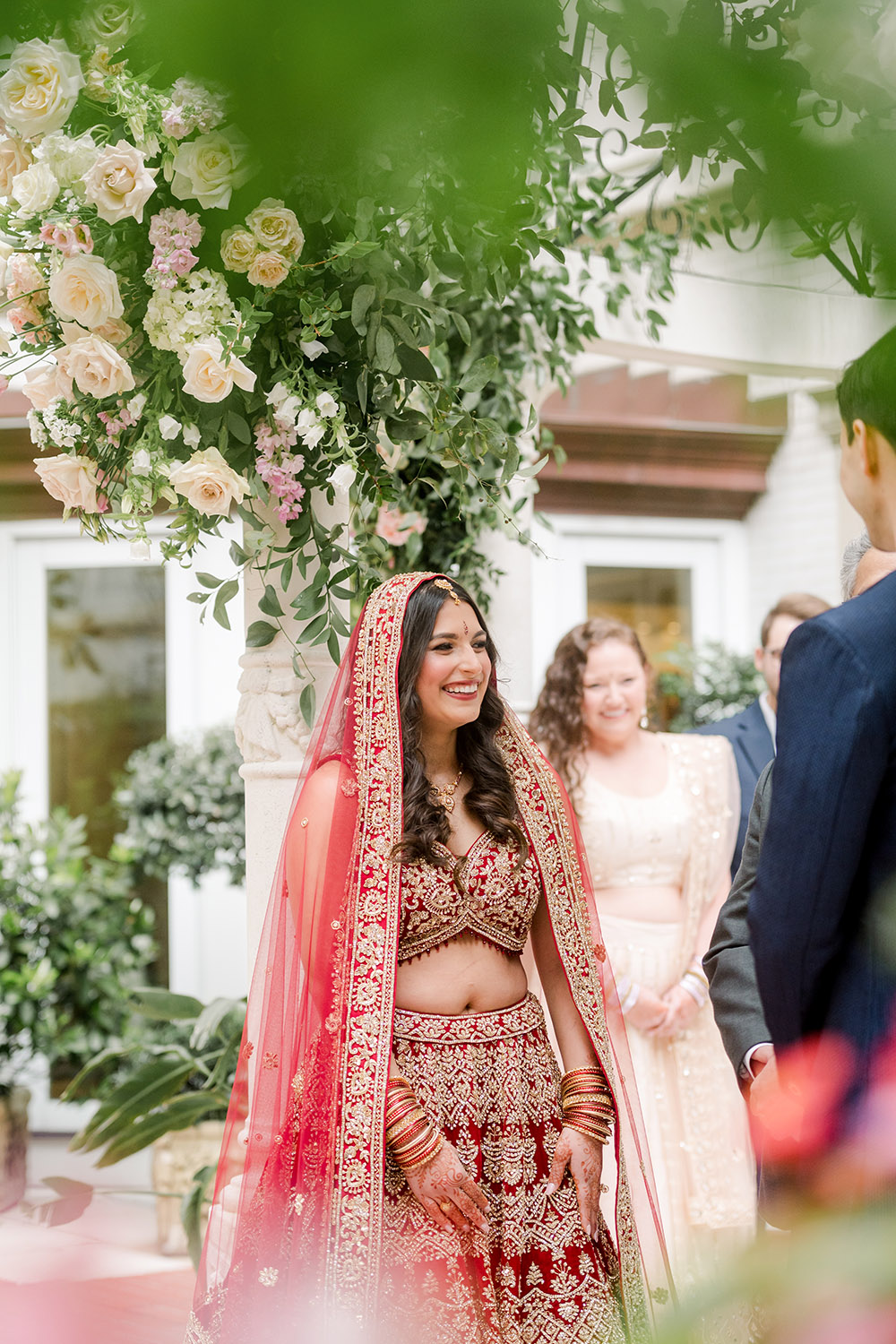 An Indian bride wearing a red and gold embroidered lehenga and matching dupatta smiles during the wedding ceremony under a floral mandap filled with roses and greenery. She stands facing the groom with guests watching behind her