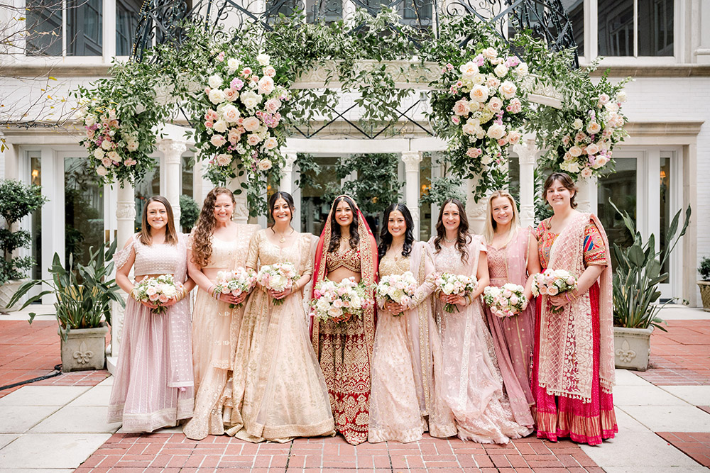 Bride dressed in a red and gold embroidered lehenga stands with her bridesmaids, who wear coordinated pastel lehengas in blush and champagne tones, each holding soft pink and white bouquets. They pose beneath a lush floral arch of roses and greenery in an elegant outdoor courtyard.