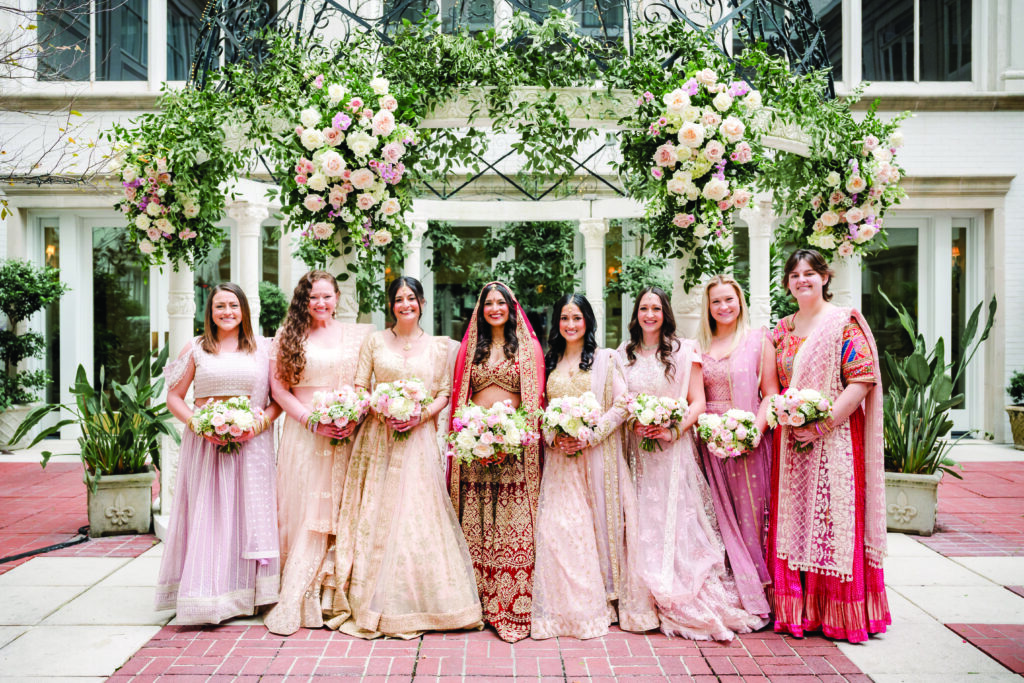 Bride dressed in a red and gold embroidered lehenga stands with her bridesmaids, who wear coordinated pastel lehengas in blush and champagne tones, each holding soft pink and white bouquets. They pose beneath a lush floral arch of roses and greenery in an elegant outdoor courtyard.