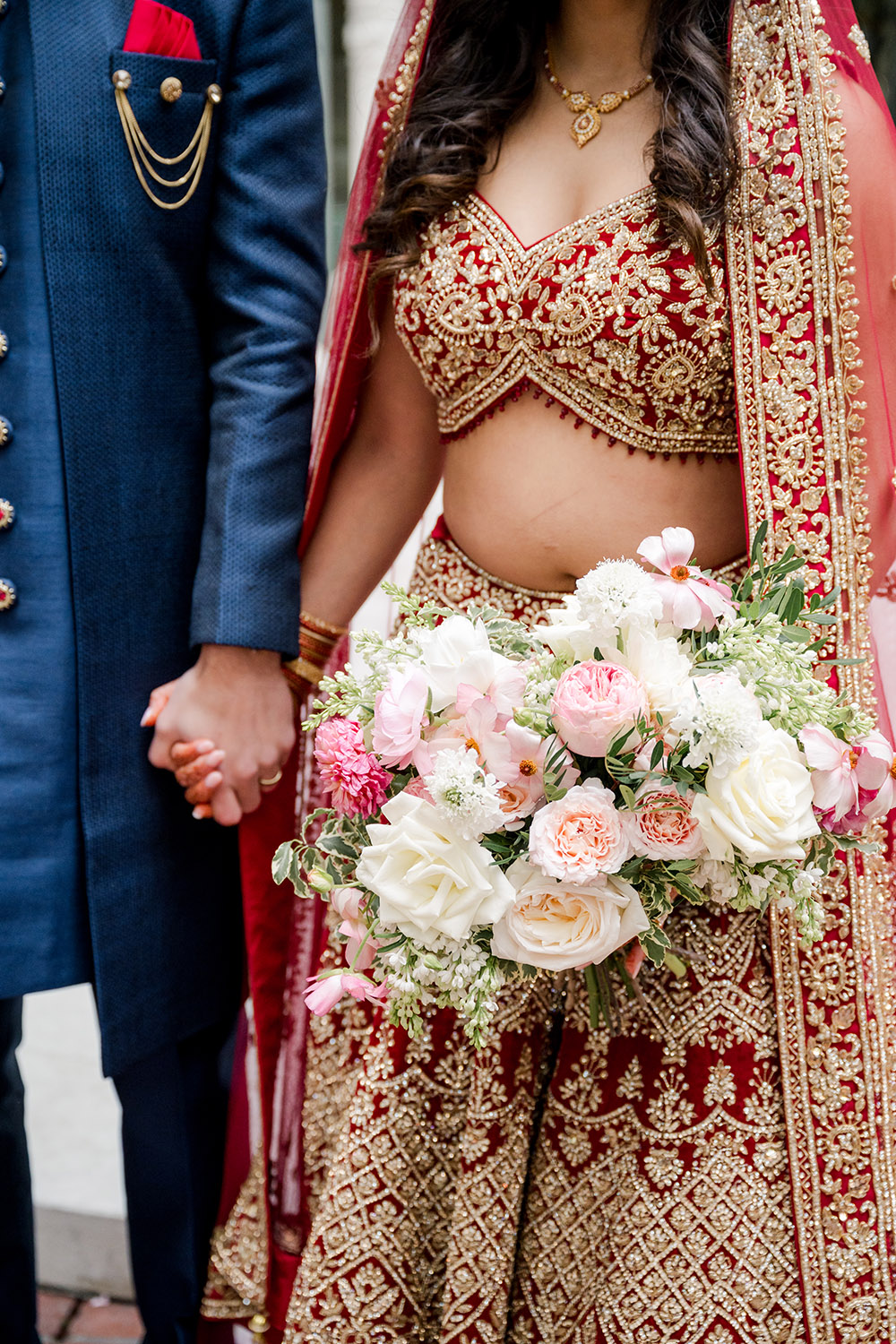 Close-up of a bride in a red and gold embroidered lehenga holding a lush bouquet of white and blush flowers with greenery. Her detailed blouse and dupatta shimmer with intricate beadwork, while she and the groom—dressed in a navy sherwani—hold hands just out of frame.