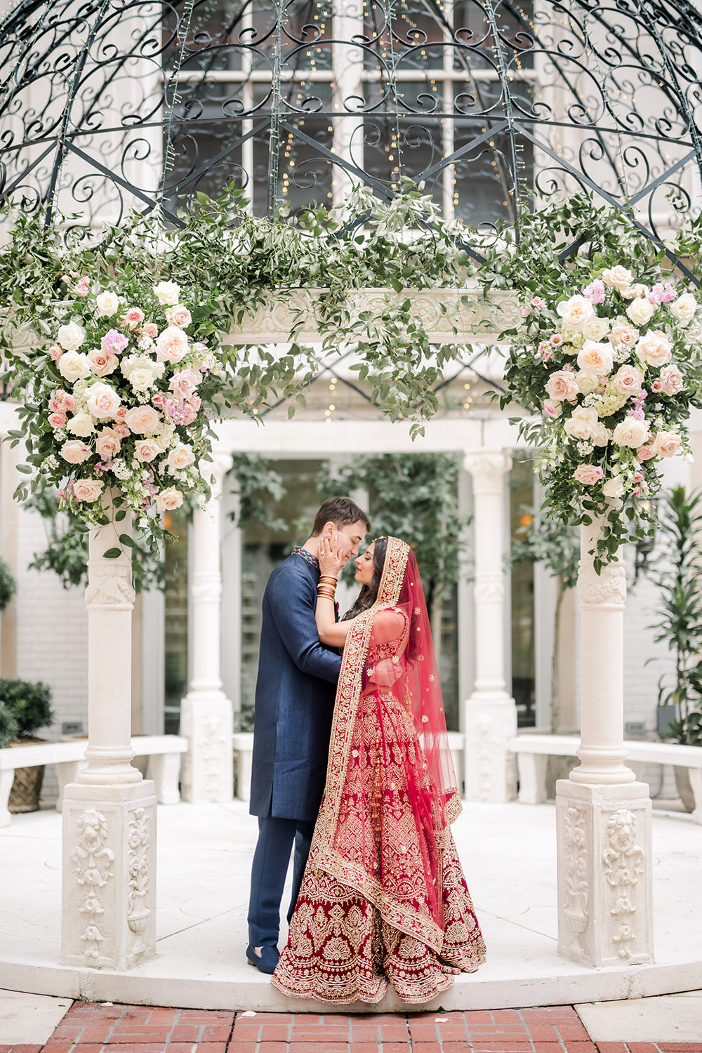 Bride and groom share an intimate kiss beneath a floral mandap adorned with roses and greenery. The bride wears a red and gold embroidered lehenga with a matching veil, while the groom wears a navy blue sherwani. They stand embraced under the ornate dome structure in the courtyard