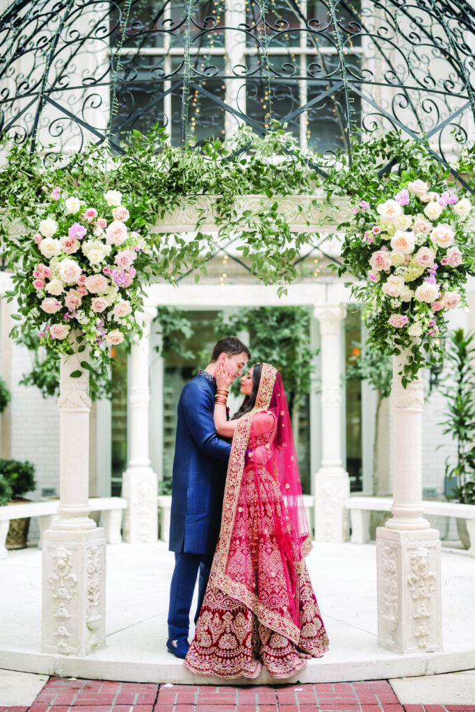 Here’s a clear, detailed, and romantic alt text for this image:

**Alt Text:**
“Bride and groom share an intimate kiss beneath a floral mandap adorned with roses and greenery. The bride wears a red and gold embroidered lehenga with a matching veil, while the groom wears a navy blue sherwani. They stand embraced under the ornate dome structure in the courtyard.”
