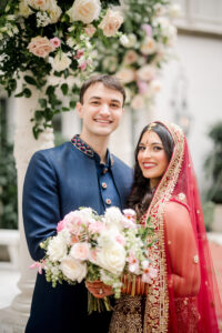 Bride and groom dressed in traditional Indian wedding attire smile beneath a floral mandap. The bride wears a red embroidered lehenga with a gold veil and holds a pastel bouquet, while the groom wears a navy blue sherwani with embroidered details.