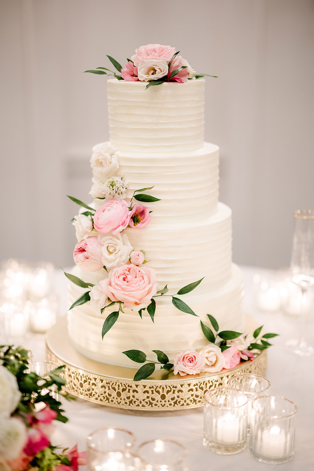 Four-tier white wedding cake with textured icing, decorated with pink and white roses and greenery cascading down the side, displayed on a gold ornate cake stand surrounded by glowing votive candles