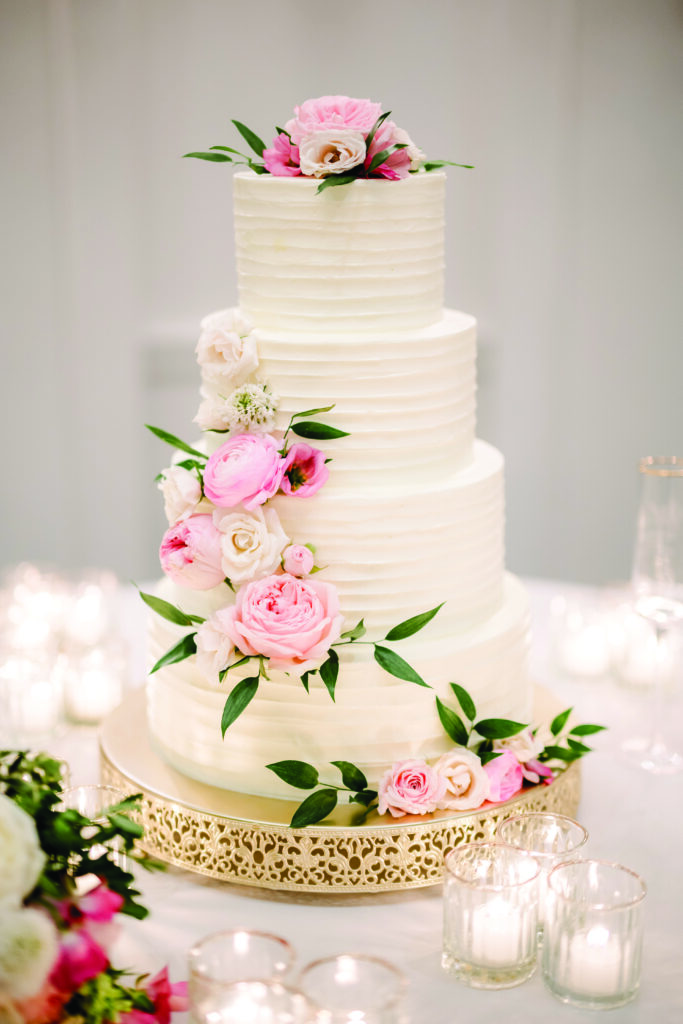 Here’s a clear, elegant, and descriptive alt text for this wedding cake image:

**Alt Text:**
“Four-tier white wedding cake with textured icing, decorated with pink and white roses and greenery cascading down the side, displayed on a gold ornate cake stand surrounded by glowing votive candles.”
