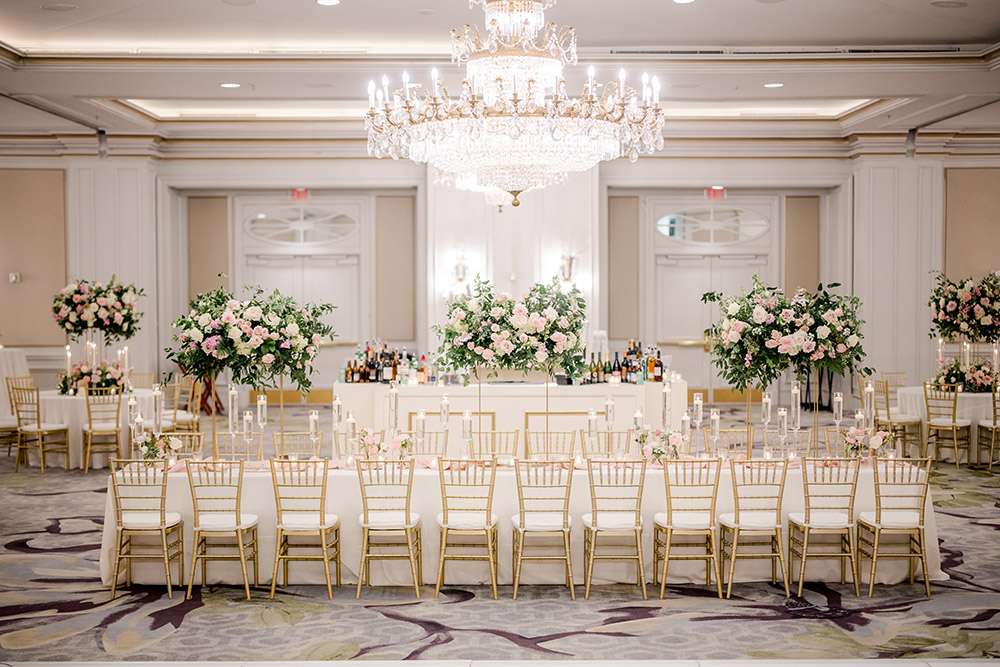 Grand wedding reception ballroom featuring a long tablescape with gold chiavari chairs, tall floral centerpieces of pink and white roses with greenery, and rows of floating candles. A large crystal chandelier hangs above, and a white bar with lush floral arrangements stands at the back of the room