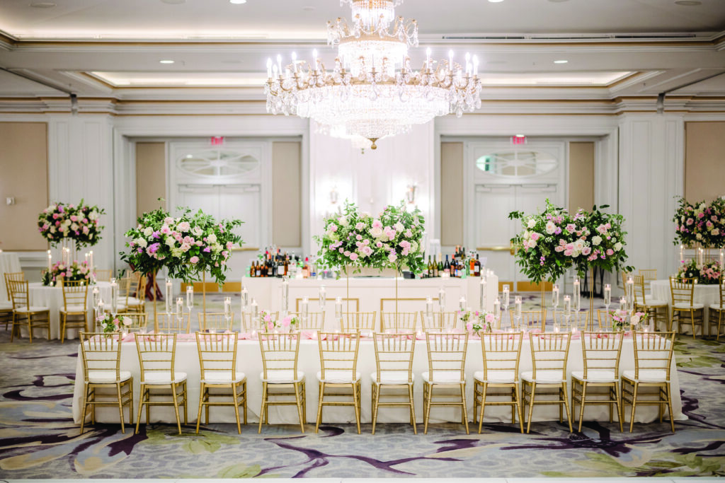 Here is a clear, elegant, and detailed alt text for this reception image:

**Alt Text:**
“Grand wedding reception ballroom featuring a long tablescape with gold chiavari chairs, tall floral centerpieces of pink and white roses with greenery, and rows of floating candles. A large crystal chandelier hangs above, and a white bar with lush floral arrangements stands at the back of the room.”
