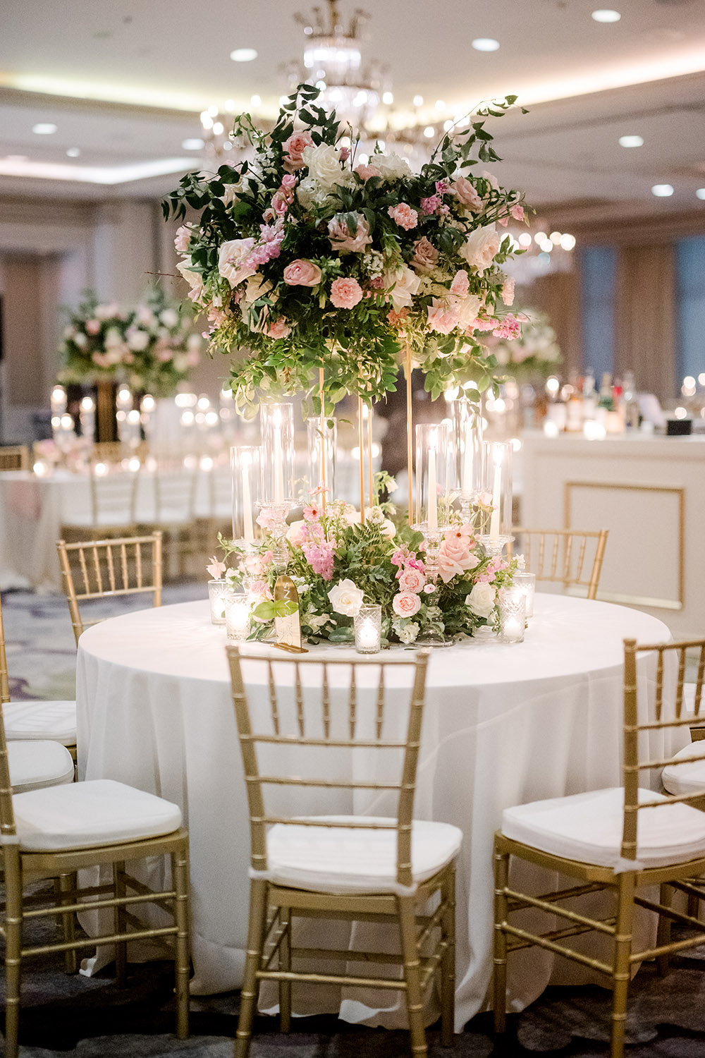 Round wedding reception table with gold chiavari chairs, featuring a tall floral centerpiece of pink and white roses with lush greenery. Clear cylinder candles surround the base of the arrangement, creating a romantic, glowing tablescape under the ballroom chandeliers.