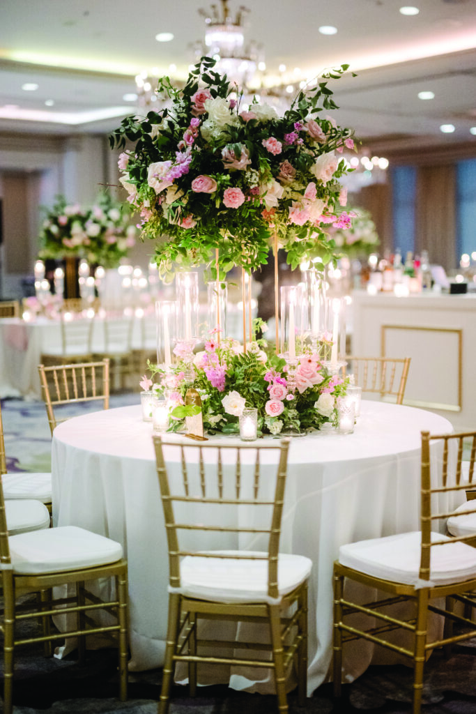 Here’s a clear and elegant alt text for this reception detail image:

**Alt Text:**
“Round wedding reception table with gold chiavari chairs, featuring a tall floral centerpiece of pink and white roses with lush greenery. Clear cylinder candles surround the base of the arrangement, creating a romantic, glowing tablescape under the ballroom chandeliers.”
