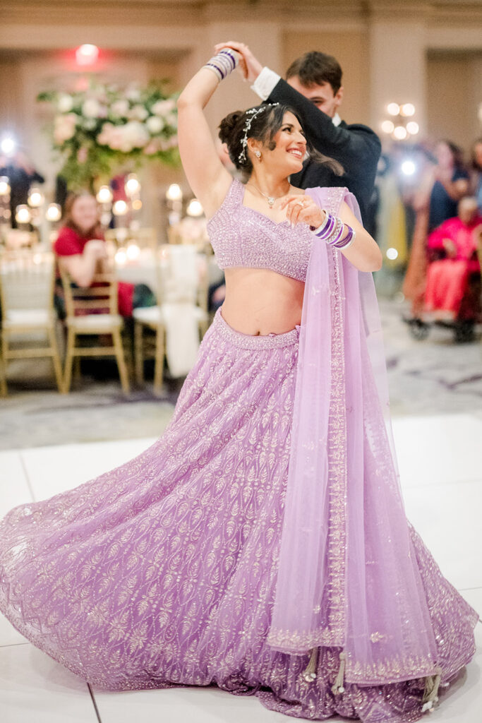 Bride twirls on the dance floor in a sparkling lavender lehenga as her groom spins her during their reception. She smiles brightly, her bangles and dupatta flowing with the movement while guests watch in the softly lit ballroom