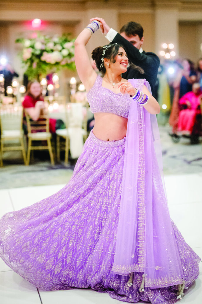 Here’s a clear, joyful, and descriptive alt text for this image:

**Alt Text:**
“Bride twirls on the dance floor in a sparkling lavender lehenga as her groom spins her during their reception. She smiles brightly, her bangles and dupatta flowing with the movement while guests watch in the softly lit ballroom.”
