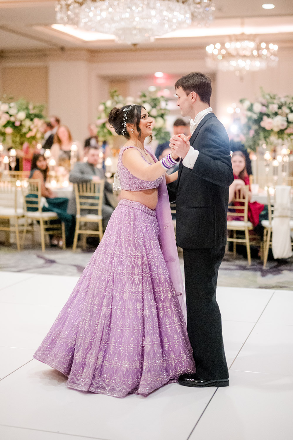Bride and groom share their first dance at the reception. The bride wears a sparkling lavender lehenga with matching bangles and a jeweled hairpiece, smiling at her groom in a black tuxedo as guests watch in the background