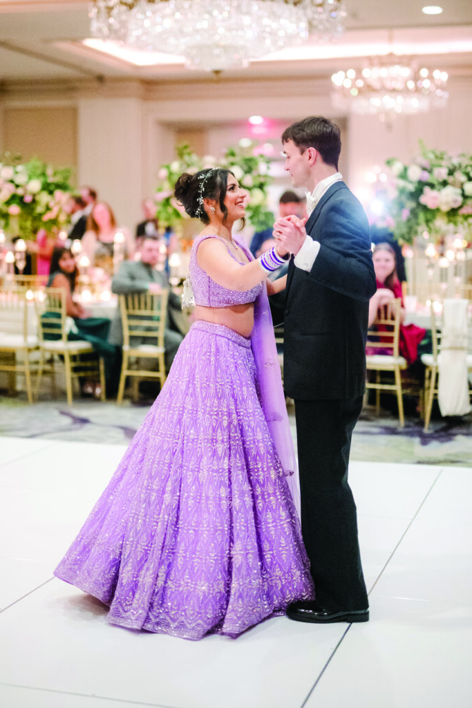 Here’s a clear, warm, and descriptive alt text for this image:

**Alt Text:**
“Bride and groom share their first dance at the reception. The bride wears a sparkling lavender lehenga with matching bangles and a jeweled hairpiece, smiling at her groom in a black tuxedo as guests watch in the background.”
