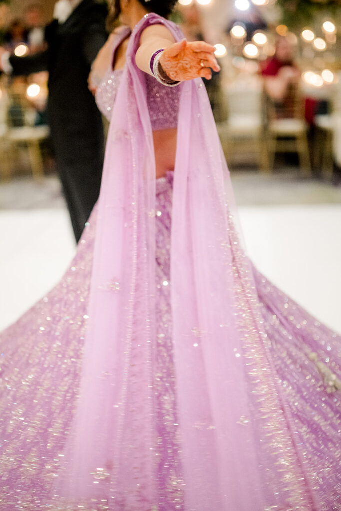 Close-up motion shot of a bride twirling in a sparkling lavender lehenga, her outstretched arm showing henna and matching bangles as the skirt swirls across the dance floor.