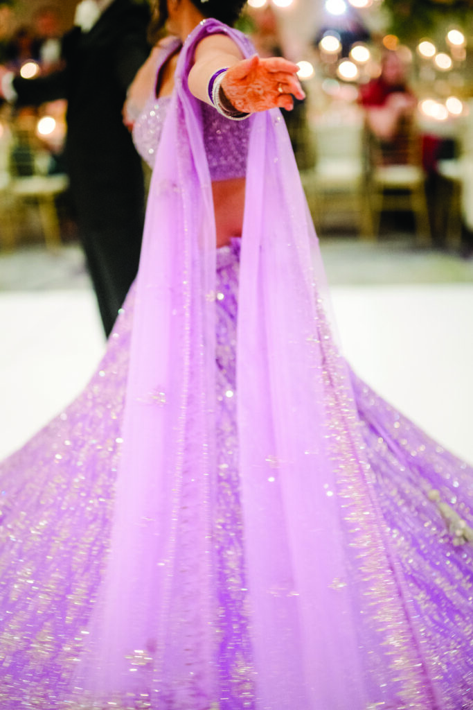 Here’s a clear, elegant alt text for this artistic, motion-filled image:

**Alt Text:**
“Close-up motion shot of a bride twirling in a sparkling lavender lehenga, her outstretched arm showing henna and matching bangles as the skirt swirls across the dance floor.”
