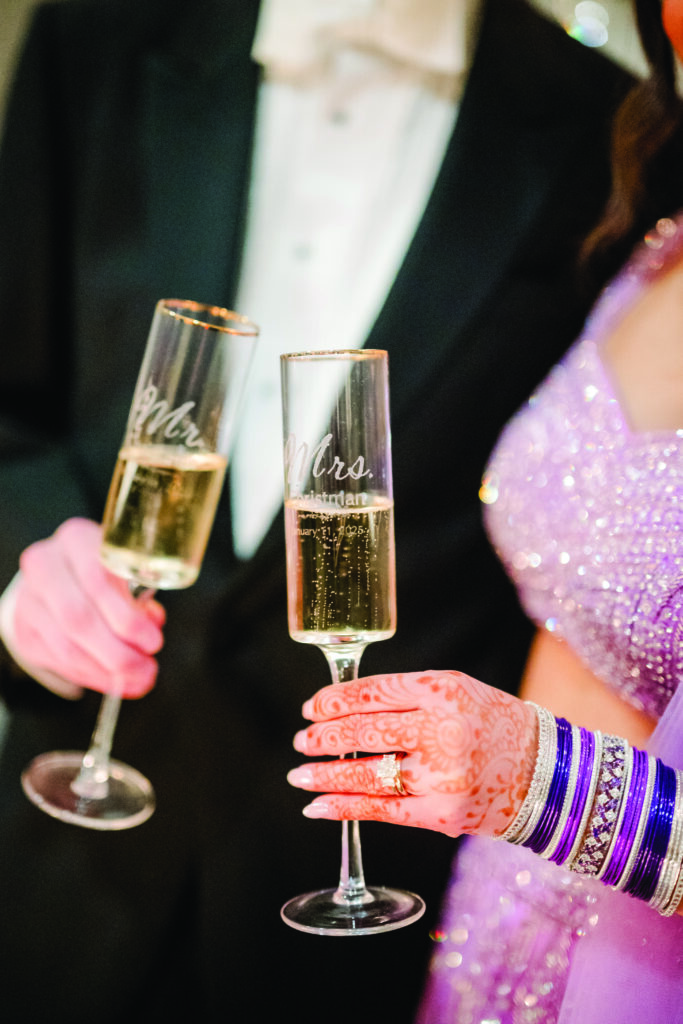 Here’s a clear, detailed, and elegant alt text for this image:

**Alt Text:**
“Close-up of the bride and groom holding personalized champagne flutes labeled ‘Mr.’ and ‘Mrs.’ during their wedding reception. The bride’s henna-covered hand, purple bangles, and engagement ring are in focus beside her sparkling lavender lehenga.”
