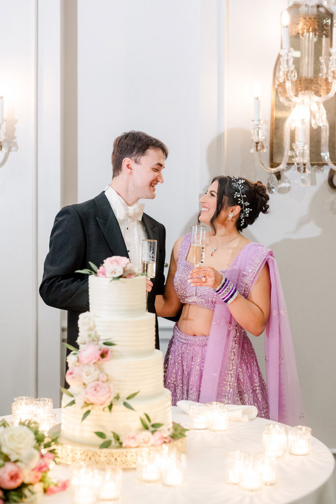 Bride and groom in a ballroom toast with champagne beside their elegant tiered wedding cake decorated with pink roses and greenery, the bride wearing a sparkly lavender lehenga and the groom in a classic black tux.