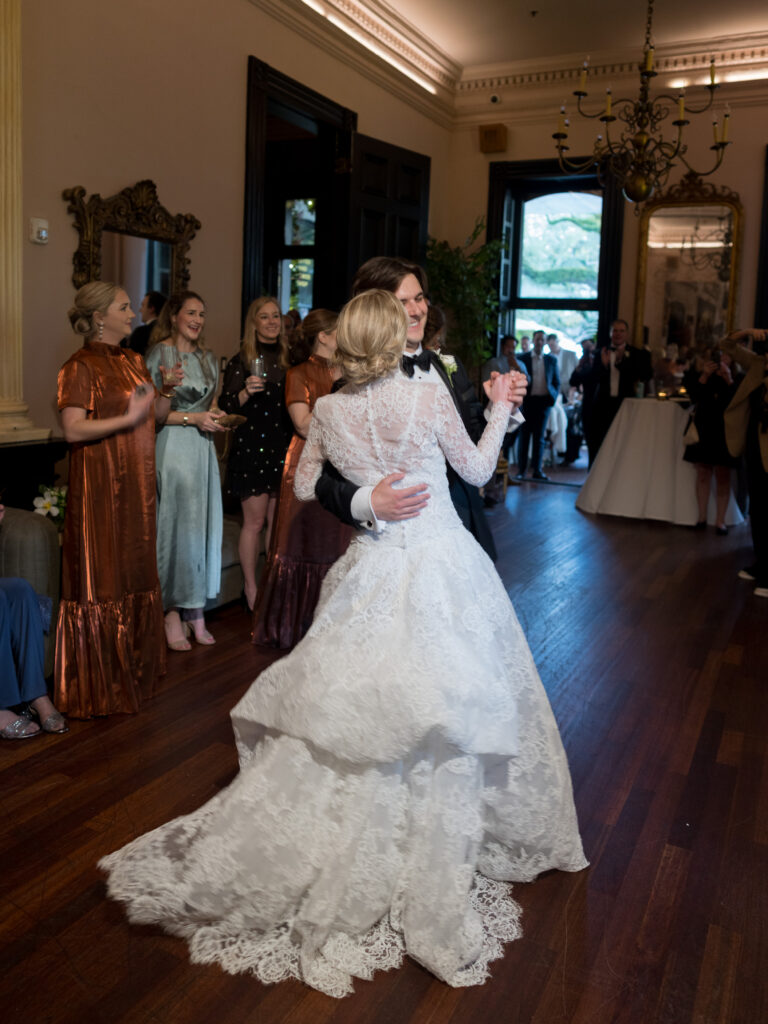 The bride and groom share their first dance in an elegant reception room, the bride’s lace gown sweeping across the wooden floor as guests smile and watch from the sidelines. Warm lighting, ornate mirrors, and tall windows create a refined, celebratory atmosphere.