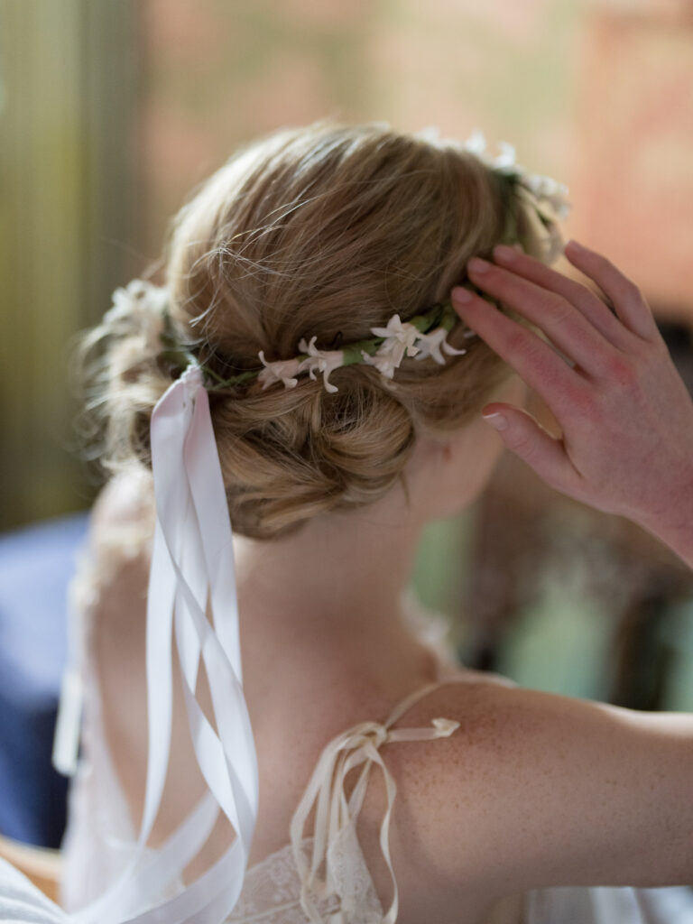 Close-up of a bride’s hairstyle from behind, featuring a soft, romantic updo adorned with a delicate floral crown and long white ribbon ties. The bride’s hand gently touches her hair as she gets ready, with soft natural light creating an intimate and elegant bridal detail shot.