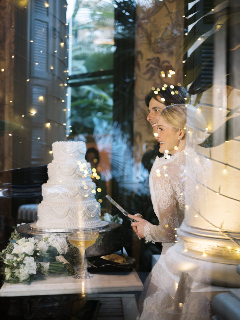 The bride and groom smile as they prepare to cut their tiered white wedding cake, seen through a reflective window that overlays soft string lights and greenery. The bride holds a cake knife beside a bouquet of white flowers and a coupe glass, creating a romantic, glowing scene.