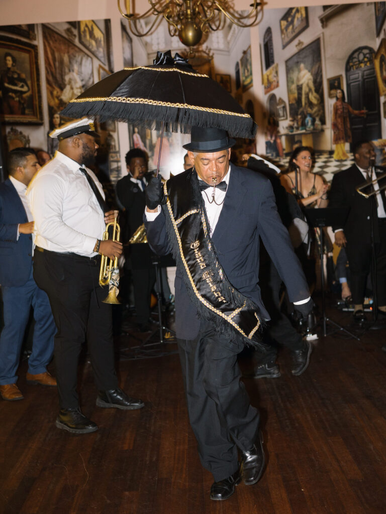 A second-line grand marshal leads the celebration, dressed in a dark suit with a gold-trimmed sash and carrying a black parasol with gold fringe. He dances while blowing a whistle, followed by a live brass band and cheering guests in the background.