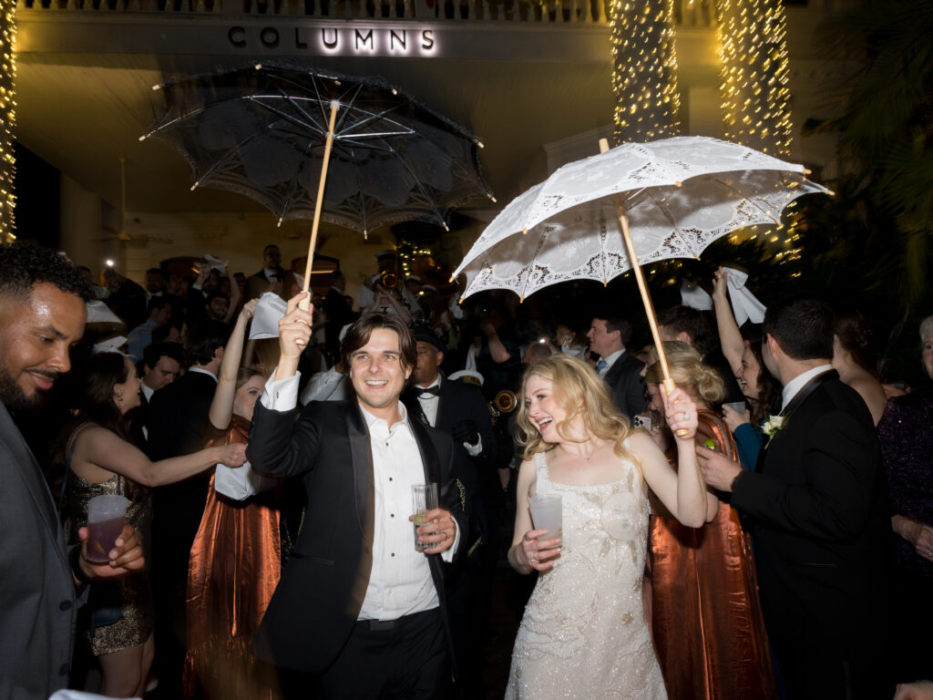The bride and groom lead a joyful second-line parade at night, each holding decorative umbrellas—his black and hers white lace—while guests wave handkerchiefs around them. The couple smiles as they dance through the crowd outside The Columns, surrounded by sparkling string lights and lively celebration.