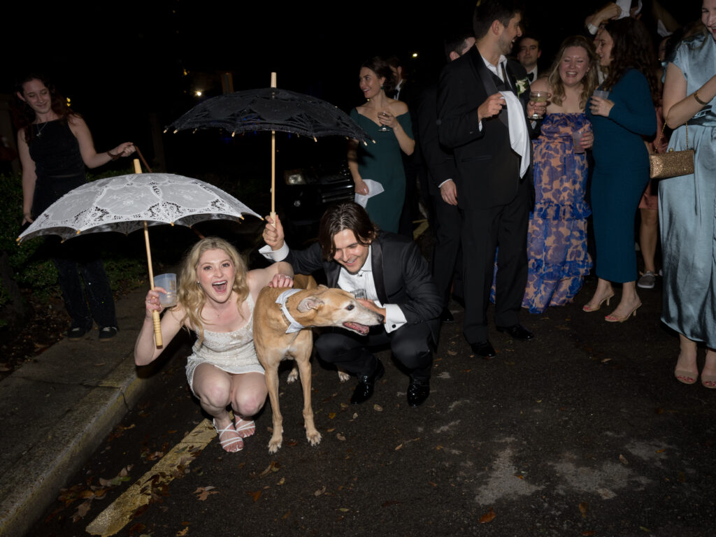 A joyful bride in a short white dress crouches beside the groom and their dog during a nighttime wedding celebration. The bride holds a drink and a white lace parasol, while the groom holds a black lace parasol and pets their dog, who is wearing a bandana. A lively group of guests in colorful attire cheer and laugh in the background on the street.