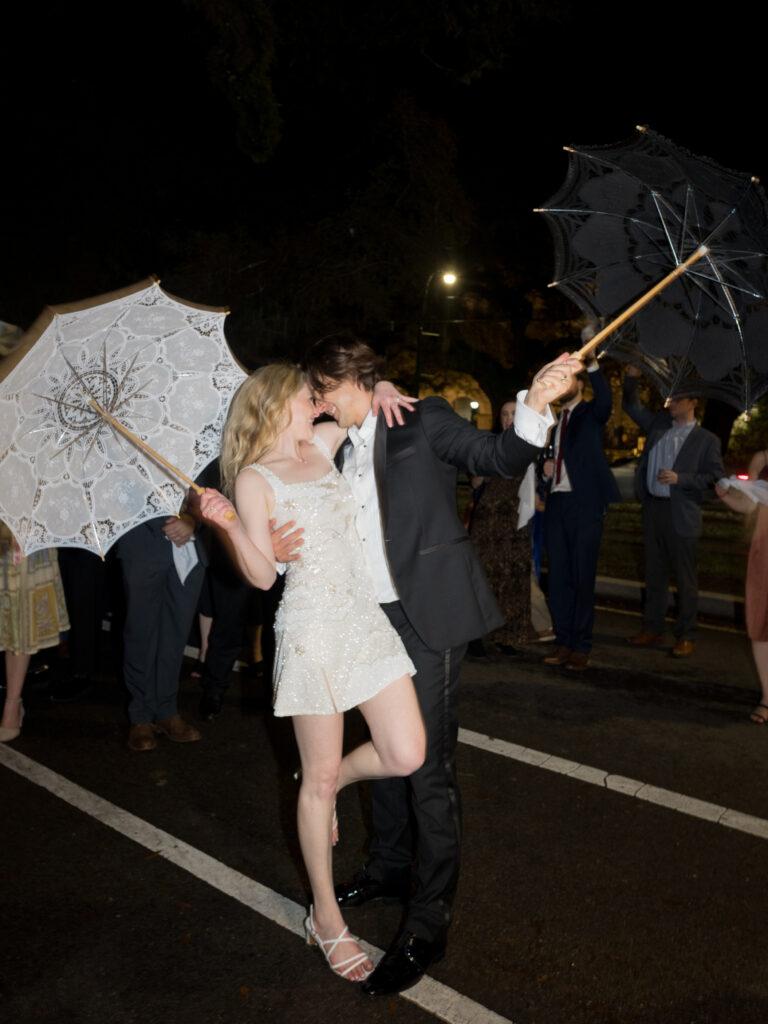 A bride in a sparkly short white dress and strappy heels shares a playful kiss with the groom during a nighttime wedding celebration. They each hold lace parasols—hers white and his black—raised above them as guests cheer in the background on the street.