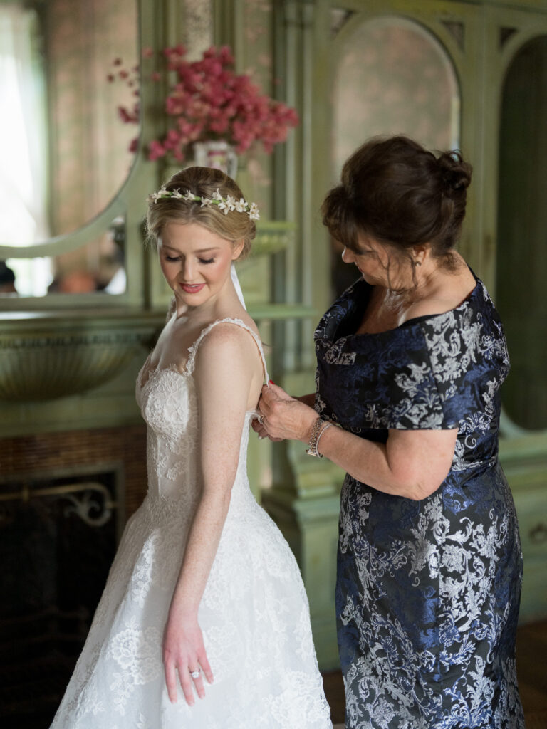 Bride stands gracefully as her mother helps fasten the straps of her lace wedding gown. The bride wears a delicate floral headpiece and looks down with a soft smile. Her mother is dressed in a navy and silver brocade gown. They are in an elegant, vintage-style room with green woodwork and soft pink flowers, capturing a tender getting-ready moment.