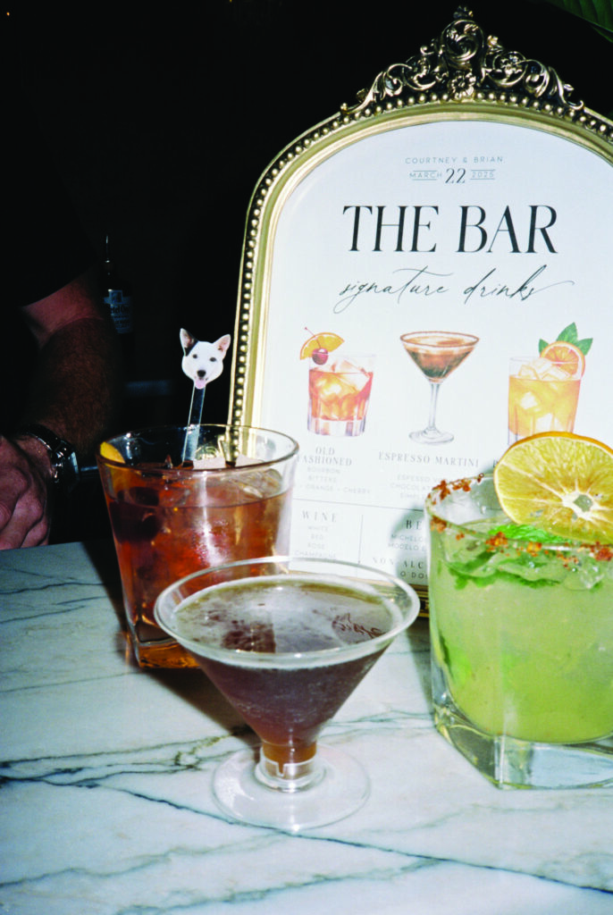 Close-up of signature wedding cocktails on a marble bar, including an Old Fashioned with a custom dog drink stirrer, an espresso martini, and a margarita, displayed in front of a framed “The Bar” signature drink menu.