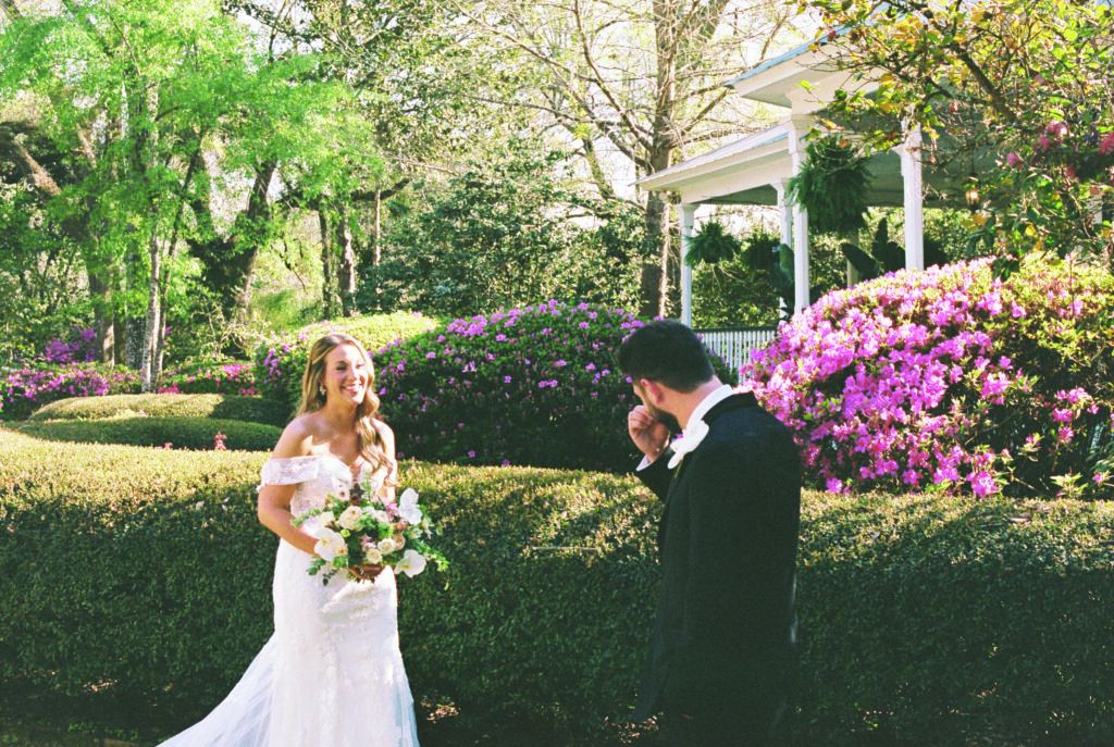 Groom wipes away tears during an emotional first look with his bride in a blooming garden, surrounded by bright pink azaleas and lush greenery as she smiles holding her bouquet.
