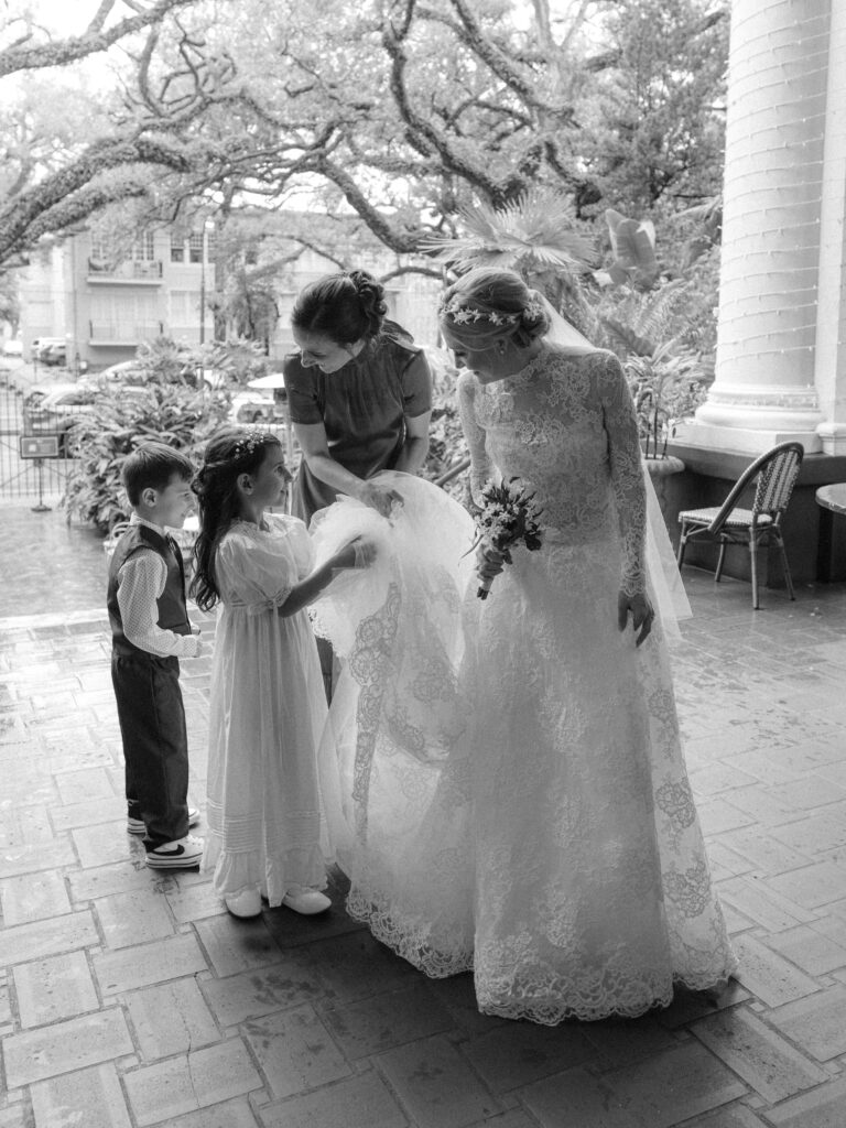 Black-and-white photo of a bride in a long-sleeve lace gown standing on a porch as a bridesmaid and two young children help arrange her dress and veil. The flower girl gently holds the lace hem while the ring bearer stands nearby. Large oak trees and historic homes create a soft, timeless backdrop for this candid pre-ceremony moment.