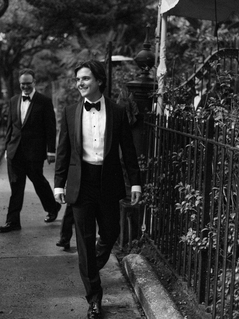 A groom in a classic black tuxedo walks down a garden-lined sidewalk, smiling and relaxed, with another man in a tuxedo following behind him in the background of the black-and-white photo.