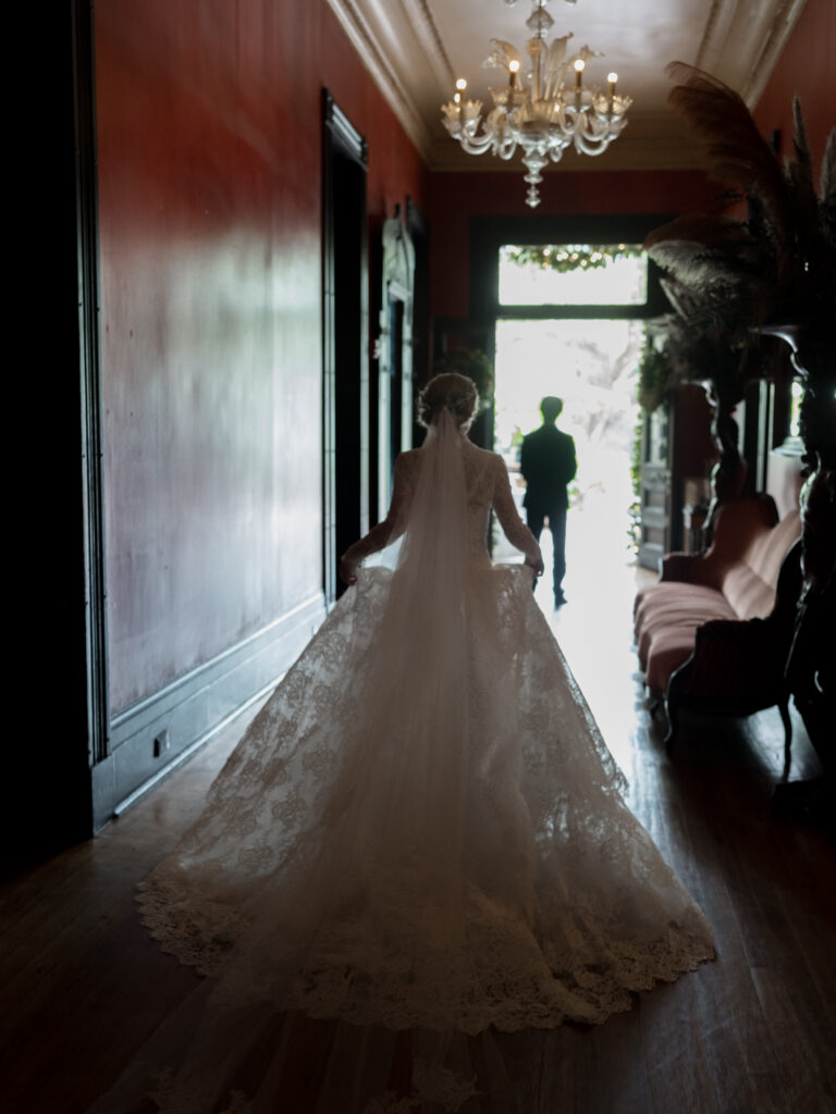 A bride in a long, lace wedding gown walks down a dimly lit, elegant hallway with dark red walls and a chandelier above, her voluminous train trailing behind her as she approaches a bright doorway where her groom waits in silhouette.