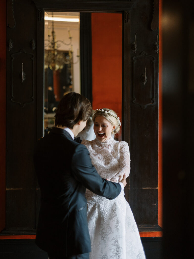 A joyful bride in a high-neck lace wedding gown laughs with excitement as she holds hands with her groom during an emotional first-look moment. They stand framed by a tall dark wooden doorway with warm reddish walls behind them, the bride’s floral headpiece and expressive smile capturing the tenderness of the scene.