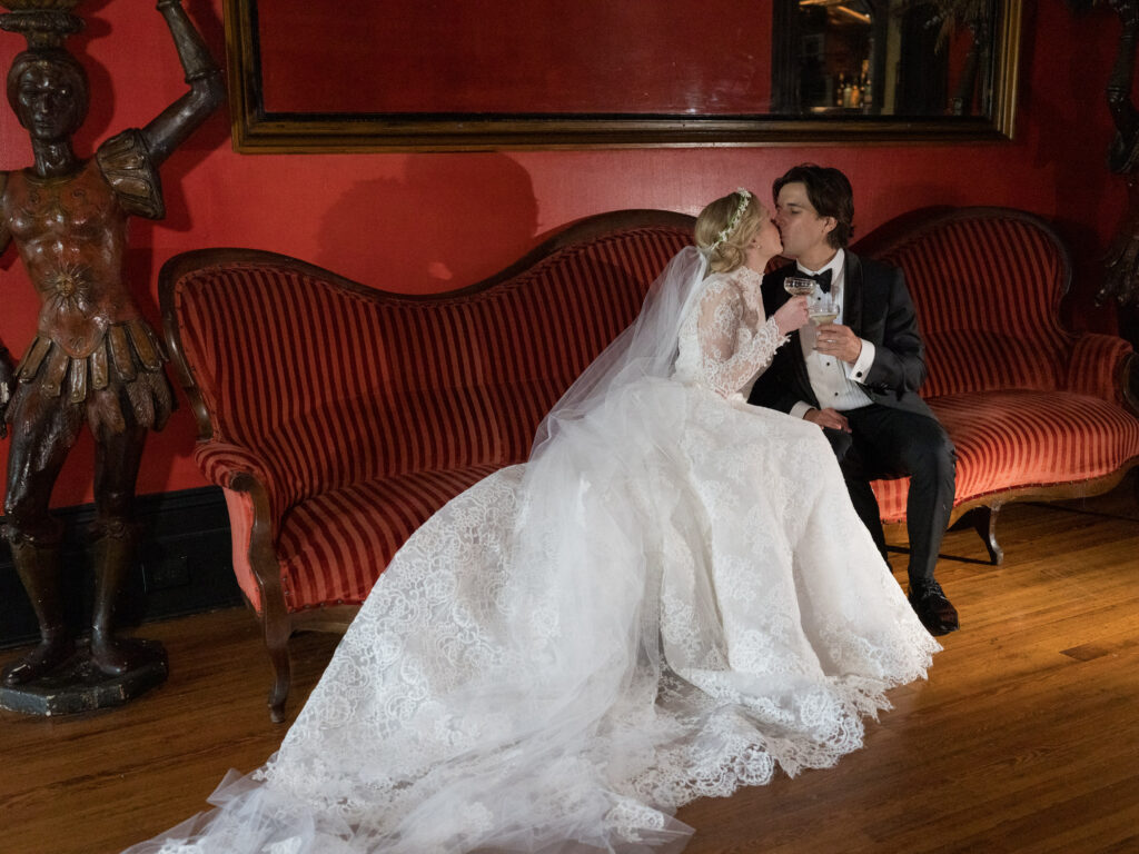 A bride in a long-sleeved lace gown and cathedral veil shares a kiss with her groom as they sit on a vintage red-striped sofa against a bold red wall. The bride’s full lace train cascades across the wooden floor while the groom, dressed in a classic black tuxedo, holds a cocktail glass. Ornate warrior-style statues frame the scene, adding dramatic character to the intimate moment.