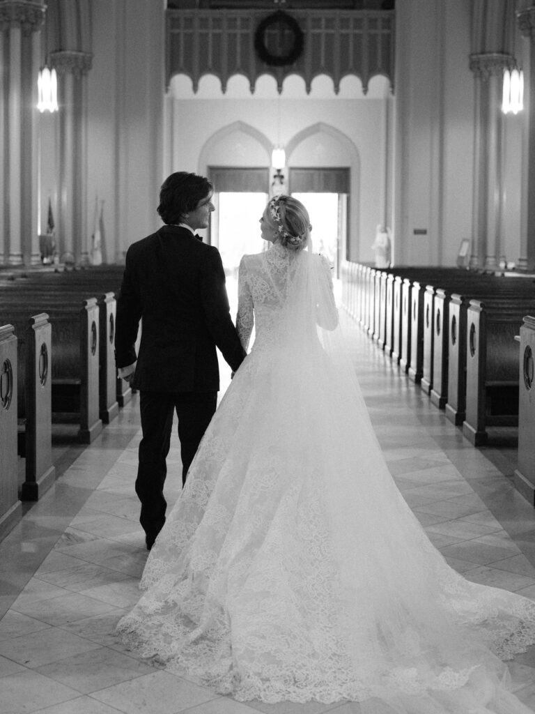 A bride and groom walk hand in hand down the aisle of a grand church, captured from behind in black and white. The bride’s long lace gown and veil flow across the marble floor as they look lovingly at each other, surrounded by tall columns and empty wooden pews.
