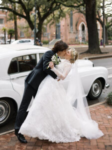 A groom in a classic black suit dips his bride for a kiss beside a vintage white Rolls-Royce. The bride wears a long-sleeve lace wedding gown, a cathedral veil, and a floral headpiece while holding a small bouquet of white flowers. They stand on a brick sidewalk under large oak trees with a historic church in the background, creating a romantic and timeless New Orleans wedding moment.