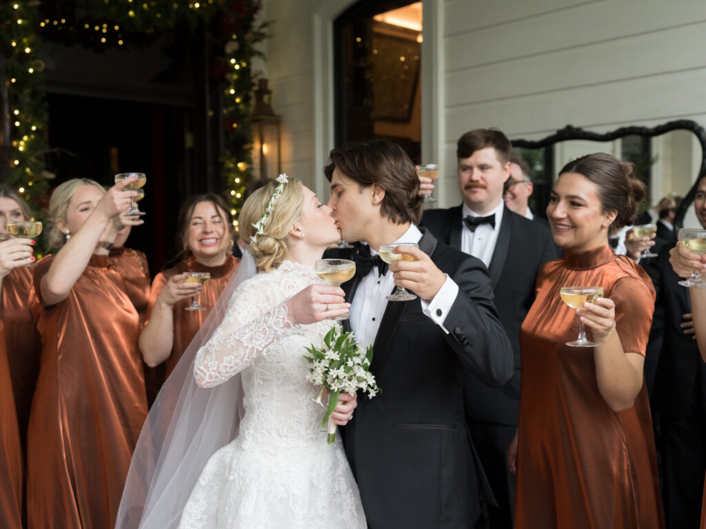 Bride and groom share a kiss while holding champagne coupes, surrounded by their wedding party raising glasses in celebration. The bride wears a lace long-sleeve gown and veil with a floral headpiece, and the bridesmaids wear satin rust-colored dresses. The groom and groomsmen are in black tuxedos. Twinkling lights and greenery frame the entrance behind them, creating a joyful and festive wedding moment.