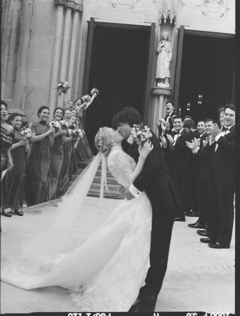 A groom dips and kisses his bride outside a grand church entrance as their wedding party lines the steps, cheering and holding bouquets. The bride’s long lace gown and flowing veil sweep across the ground, while bridesmaids and groomsmen celebrate around them in this joyful black-and-white moment.