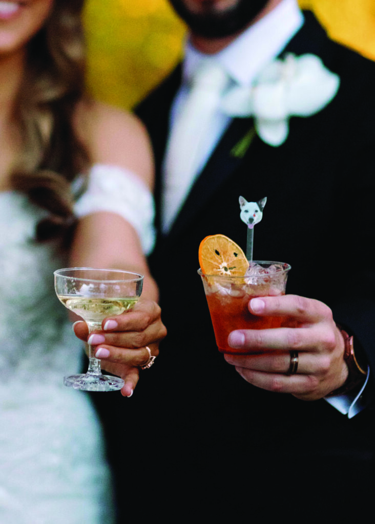 Close-up of bride and groom holding their signature wedding cocktails, including a champagne coupe and a custom drink with a dog-shaped stir stick and dried citrus garnish, celebrating in elegant wedding attire.