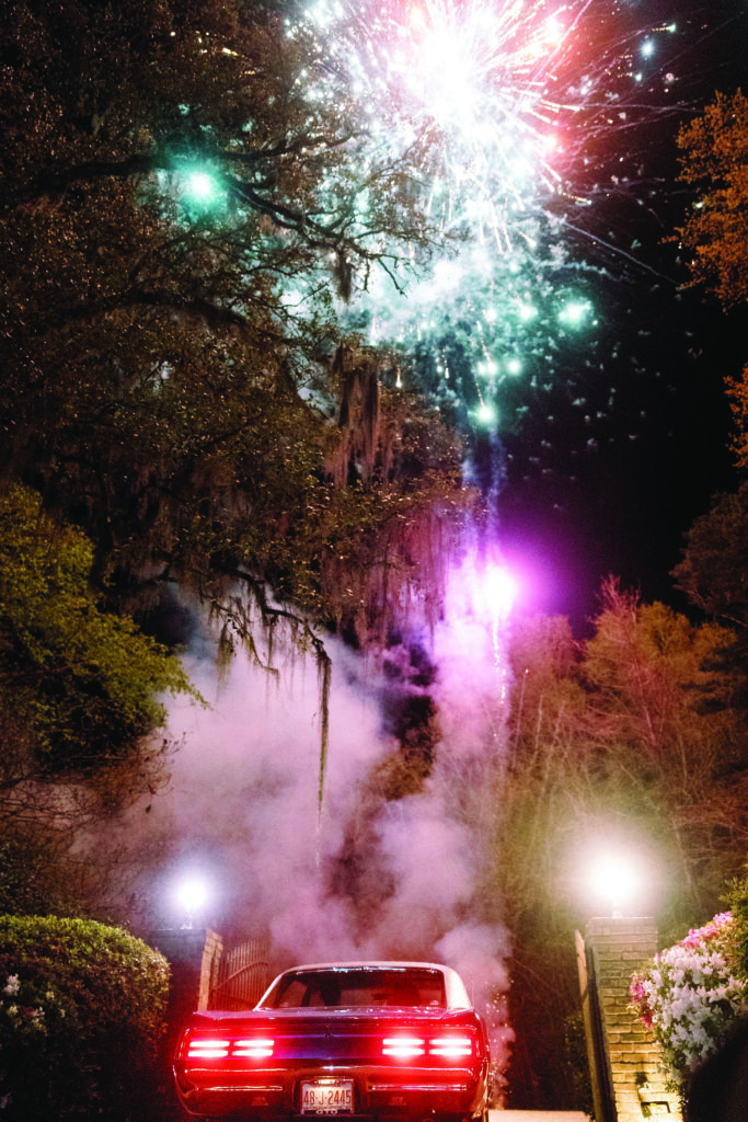 A classic red car drives away under a sky full of bright fireworks, creating a dramatic and celebratory nighttime wedding exit scene with smoke and glowing lights along the driveway.