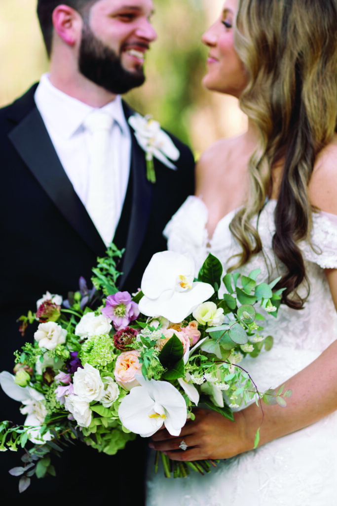 Close-up of a bride and groom smiling at each other while the bride holds a lush bouquet of white orchids, roses, and greenery, with the groom in a black tuxedo and the bride in an off-the-shoulder lace gown.