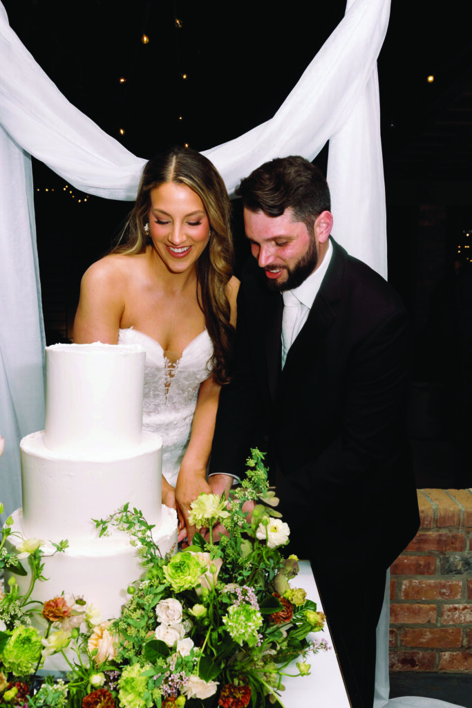 A groom in a black suit carefully pours champagne into a cascading tower of coupe glasses while the bride, wearing a strapless white wedding gown, smiles beside him during an outdoor wedding celebration surrounded by warm golden light and lush greenery.