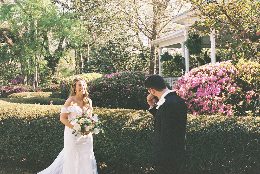 Groom wipes away tears during an emotional first look with his bride in a blooming garden, surrounded by bright pink azaleas and lush greenery as she smiles holding her bouquet.